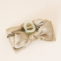 The Darling Effect’s Green Satin Bow Claw Clip secures a beige satin bow, creating an elegant updo accessory. Displayed on a light, textured surface, the translucent clip adds a soft green accent to your hairstyle.
