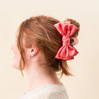A woman with light brown hair in an updo, secured by The Darling Effect's Rose Satin Bow Claw Clip. She wears a white textured sweater and small hoop earrings, shown in profile against a light beige background.