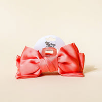 The Darling Effect’s Rose Satin Bow Claw Clip is a large, shiny coral pink hair accessory with a central knot on a clear plastic clip, shown on a light beige background with a round white tag reading "the darling effect.