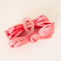 The Darling Effect's Rose Satin Bow Claw Clip features a decorative satin ribbon bow in pink, perfect for updos and use as a stylish hair accessory, shown on a light-colored surface.