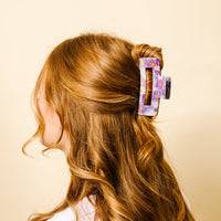 A person with long, wavy, reddish-blonde hair styled in a half-updo, secured with the-darling-effect's Bouquet Beauty Purple Claw Clip, is seen from the back against a pale yellow background.