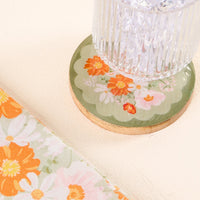 A clear glass with ice rests on the-darling-effect’s Bouquet Beauty Sage Wood Enamel Coaster, decorated with orange, pink, and white flowers. Beside it, a fabric with a matching orange floral pattern complements the coaster.