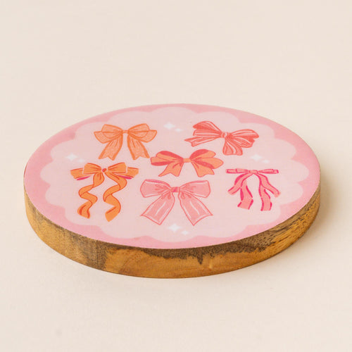 The Blushing Bows Wood Enamel Coaster by the-darling-effect features a pink scalloped edge and six orange and pink bows, combining durability and charm to protect your table in style.