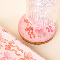 A clear glass with ice rests on the Blushing Bows Wood Enamel Coaster by the-darling-effect, featuring red and pink bow illustrations. Nearby, matching bow-patterned wrapping paper is partially visible.