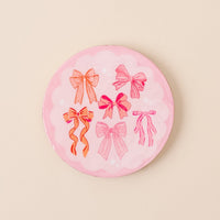 The-darling-effect’s Blushing Bows Wood Enamel Coaster is a round, pink coaster featuring six illustrated pink and red bows on a pale scalloped background—a charming and durable addition to your collection.