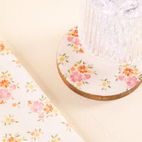 The Endless Daydream Cream Wood Enamel Coaster by the-darling-effect, with its pink and orange floral design, adds charm to any light surface, paired here with a clear glass and matching floral-patterned fabric.