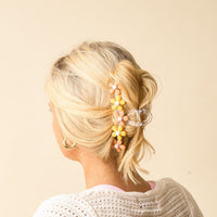 A woman with blonde hair styled in a loose updo, secured with The Darling Effect’s Peachy Daisy Claw Clip and pastel flower hair pins. She wears gold hoop earrings and a white knit top against a light beige background.