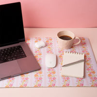 A laptop, wireless mouse, AirPods case, coffee cup, notepad, and pen are arranged on the Petal Parade Pink Style Station Desk Mat by the-darling-effect, featuring a floral pattern and non-slip base against a pink background.