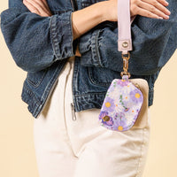 A person in a denim jacket and white pants wears the-darling-effect’s Bouquet Beauty Purple Dynamic Duo Pouch Wristlet, attached as a lavender lanyard for a stylish, hands-free accessory.