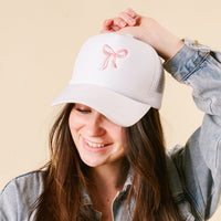 A woman in a denim jacket wears the Bow Pink Trucker Hat by the-darling-effect, smiling as she holds its brim against a beige background—showcasing how this trendy hat can elevate any look.