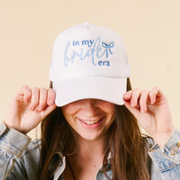 A smiling woman with long brown hair wears the-darling-effect’s In My Bride Era Trucker Hat, featuring blue embroidered lettering, and styles it with a light denim jacket for a trendy bridal look.