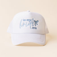 The-darling-effect’s In My Bride Era Trucker Hat is a white hat with "in my bride era" in light blue embroidery and a small blue bow by "bride," shown on a neutral background.
