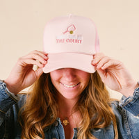 A person smiles, holding the brim of the-darling-effect’s Meet Me at the Court Trucker Hat—pink with "meet me at THE COURT," a mini tennis racket and ball—stylishly paired with a denim jacket.
