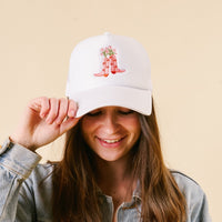 A smiling person touches the brim of the-darling-effect's Cowgirl Boots Trucker Hat, a trendy white cap featuring pink boots with flowers, against a plain beige background.