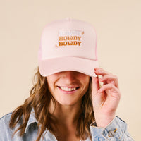 A smiling person holds the brim of the-darling-effect’s Howdy Howdy Howdy Trucker Hat—a light pink hat with colorful white, orange, and yellow embroidery—against a beige background that highlights its fun vibe.