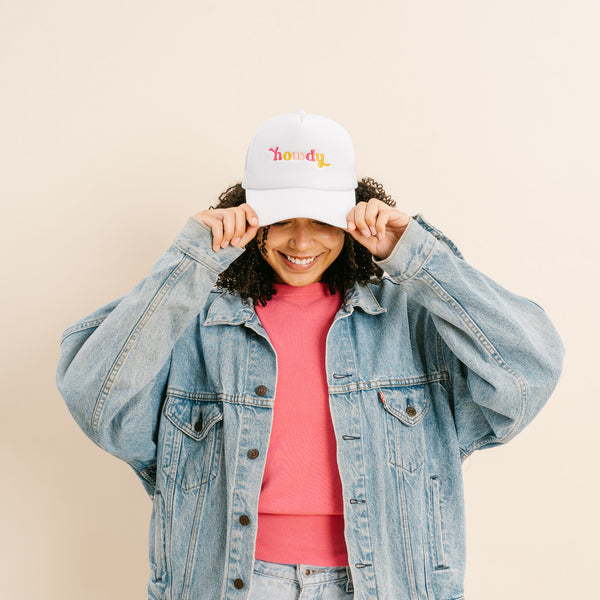 A person wearing a denim jacket, pink shirt, and The Darling Effect Howdy Trucker Hat smiles while holding the brim of the hat with both hands against a neutral background.