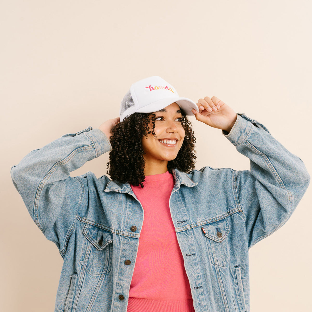A smiling person with curly hair models The Darling Effect's Howdy Trucker Hat, paired with a pink shirt and light blue denim jacket, holding the hat's brim against a beige background.