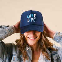 A smiling person wears the Lake Life Trucker Hat by the-darling-effect—a trendy navy blue embroidered cap—paired with a denim jacket, hands behind their head.