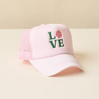 The-darling-effect LOVE Pickleball Trucker Hat in light pink mesh features "LOVE" in green embroidery with a pink strawberry for the “O.” Shown on a beige surface.