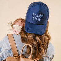 Back view of a woman wearing the-darling-effect's Mom Life Trucker Hat, featuring embroidered "MOM LIFE" and a bow, paired with a denim jacket and holding a baby in a beige headband.