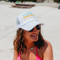 A woman in sunglasses and a pink swimsuit top smiles on a sunny beach while wearing the-darling-effect's Sunny Daze Trucker Hat, featuring colorful "sunny days" embroidery.
