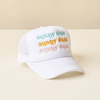 The-darling-effect’s Sunny Daze Trucker Hat features “sunny daze” embroidered three times in pastel blue, yellow, and pink on white mesh, shown atop a cream surface with a beige background.