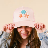 A person in a light denim jacket smiles and holds the brim of the-darling-effect’s Seashells Trucker Hat, embellished with seashells, starfish, and bead accents, standing against a cream background.