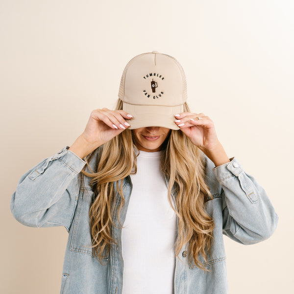 A woman in a denim jacket adjusts The Darling Effect's Tumbler Fan Club Trckr Hat—a tan trucker hat featuring an embroidered logo and tumbler cup graphic.