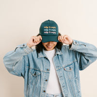 A person in a denim jacket and white t-shirt smiles while adjusting The Darling Effect’s Take it Easy Trckr Hat, featuring "take it easy" embroidered three times in colorful letters. The background is plain and light-colored.