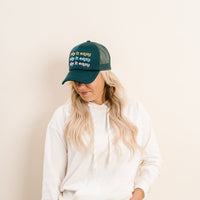 A woman with long blonde hair wears The Darling Effect's Take it Easy Trckr Hat, a trendy dark green cap with repeated embroidery, along with a white hoodie. She stands against a light background, looking down and smiling slightly.
