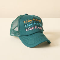 The Darling Effect’s Take it Easy Trckr Hat is a teal mesh trucker hat with “take it easy” embroidered three times in yellow, blue, and pink on the front, shown resting on a light-colored surface.