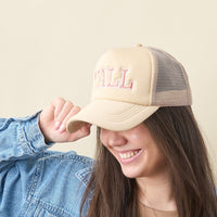 A smiling person wears the-darling-effect's Y'all Trucker Hat, featuring "TALL" in pink letters, with a blue denim jacket and holds the cap brim against a plain light background.