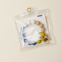 The Darling Effect’s Classic Coast Hands-Free Keychain Wristlet features a set of colorful round silicone beads on a keyring, stylishly displayed in a clear plastic pouch with snap closure on a light beige surface.
