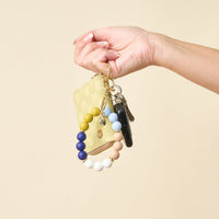 A hand holding The Darling Effect Classic Coast Hands-Free Keychain Wristlet, featuring a yellow wallet, several keys, and a silicone beaded wristlet in shades of yellow, blue, white, and beige against a plain light background.