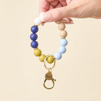 A hand holds The Darling Effect’s Classic Coast Hands-Free Keychain Wristlet, featuring round silicone beads in white, blue, tan, and green, and finished with a gold clasp adorned with a heart-shaped tag reading “the tiny acorn.”.