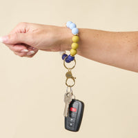 A hand wearing The Darling Effect Classic Coast Hands-Free Keychain Wristlet holds house keys and a black car key fob against a plain beige background.
