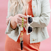 A woman wearing an orange dress and a light green jacket poses against a pink background, holding The Darling Effect Dash of Daisy Hands-Free Keychain Wristlet—featuring green beads, keys, and a car fob—in one hand along with beige sunglasses.