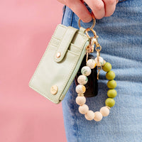 A hand holds The Darling Effect Dash of Daisy Hands-Free Keychain Wristlet in sage green, featuring a matching beaded keyring with keys and a black fob, against a pink background. The person is wearing blue jeans.