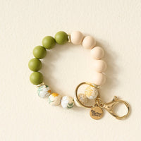 The Dash of Daisy Hands-Free Keychain Wristlet by The Darling Effect features olive green, cream, and floral-patterned silicone beads arranged in a circular shape, accented with a gold clasp and a small gold charm engraved with the brand name, displayed on a light background.