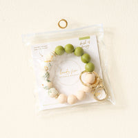 The Darling Effect Dash of Daisy Hands-Free Keychain Wristlet features green, floral, and beige silicone beads with a gold keyring and clasp, packaged in a clear zippered bag labeled “hands free.”.
