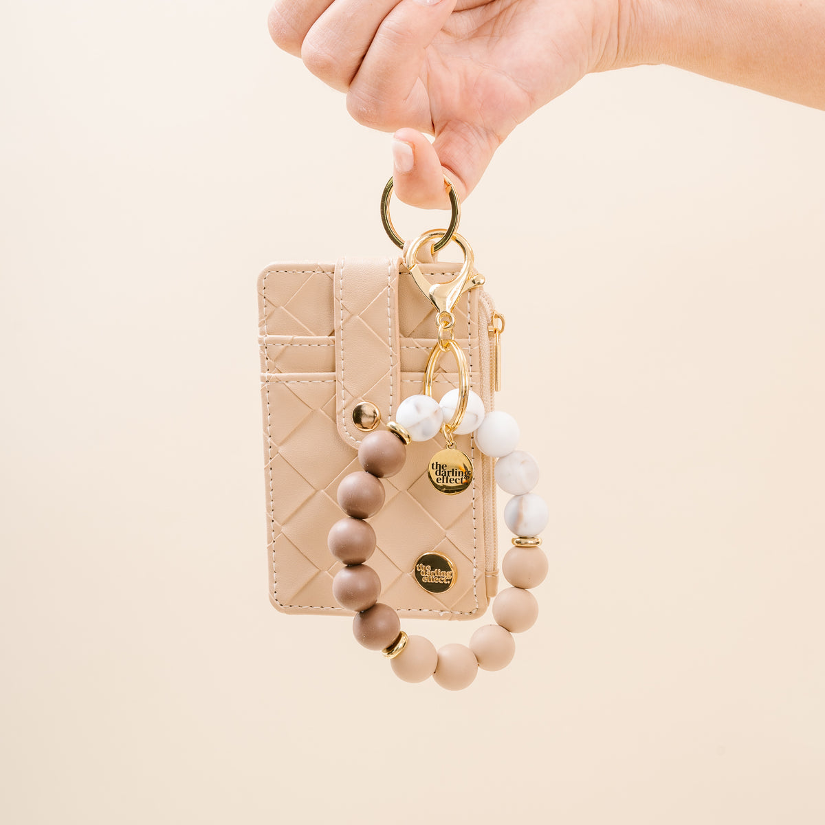 A hand holds The Darling Effect’s Hands-Free Keychain Wristlet in Mocha Swirl—a beige quilted cardholder with gold hardware, attached to a silicone bead bracelet featuring tan, brown, and white marbled beads accented with small gold charms.