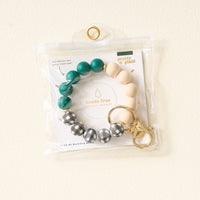 The Darling Effect's Pretty in Plaid Hands-Free Keychain Wristlet features green, beige, and black-and-white plaid silicone beads on a gold key ring and clasp, displayed in a clear resealable pouch.