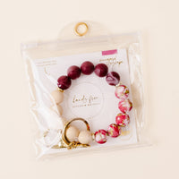 The Vineyard Escape Hands-Free Keychain Wristlet from the-darling-effect is displayed in a clear pouch, featuring large maroon, cream, and floral silicone beads. A gold keyring and clasp offer a convenient way to hold your keys. Inside, a card reads “Vineyard Escape Hands-Free Keychain Wristlet.”.