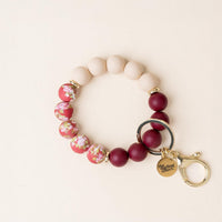 Introducing the Vineyard Escape Hands-Free Keychain Wristlet by the-darling-effect, featuring pink, maroon, and floral-patterned silicone beads with gold rings and a gold clasp. A small round charm reads ‘the Giving effect.’ The circular wristlet keychain is displayed on a cream background.