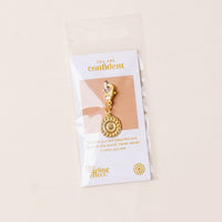 The-darling-effect Confident Jewelry Trait Charm Gold, featuring a decorative circular design, is attached to a card reading, "You are confident. Believe you are beautiful and deserve the world. Never waver in who you are.