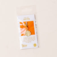The Strong Jewelry Trait Charm Gold by the-darling-effect is a gold keychain with a circular strength charm, packaged on a card that says “You are strong” with an encouraging message and displays the the-darling-effect logo at the bottom.