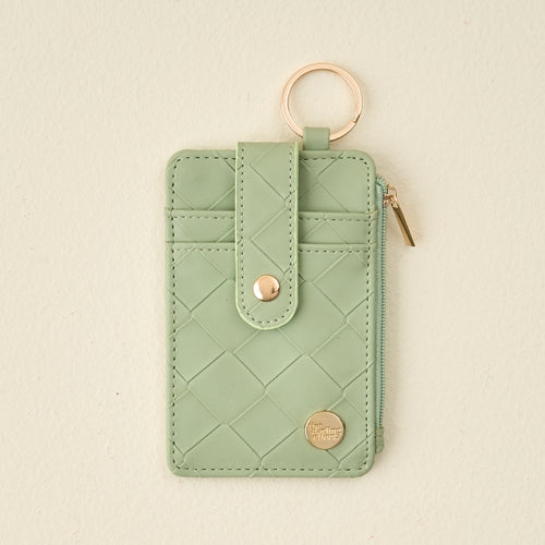 The Darling Effect's Sage Woven Keychain Wallet features a small quilted light green design, gold logo detail, snap strap, gold keyring, and zippered coin holder. Shown here against a beige background.