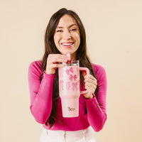 A smiling woman with long dark hair wears a bright pink top and holds the-darling-effect’s Blushing Bows 40 oz Lifestyle Flip Straw Tumbler, decorated with bows, against a beige background.