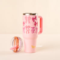 The Blushing Bows 40 oz Lifestyle Flip Straw Tumbler by the-darling-effect, in pink with bow decorations and yellow text near the base, sits on a light surface next to its leak-resistant clear flip straw lid with a pink straw.