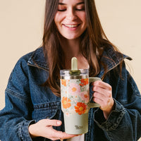 A woman in a denim jacket smiles while holding the-darling-effect’s Bouquet Beauty Sage 20 oz Lifestyle Flip Straw Tumbler against a light beige background.